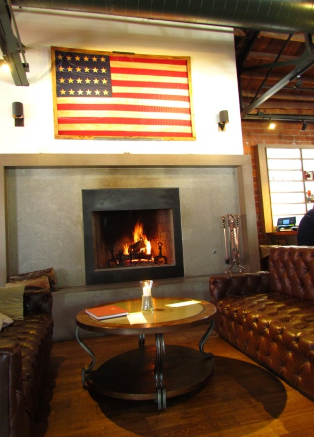 A historic American flag with 24 stars hangs above a welcoming fireplace. Guests can relax on the tufted brown leather couches that flank the round wooden table in this inviting seating area of the Third Street Social Restaurant.