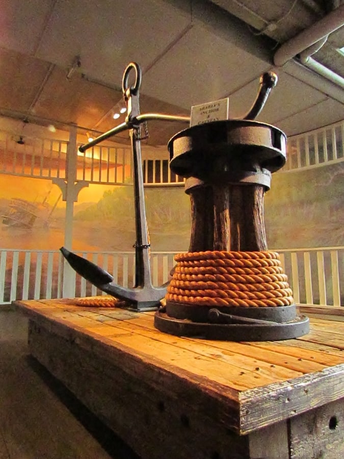 The metal anchor and wooden capstan from the Arabia Steamboat are displayed on a wooden riser. 