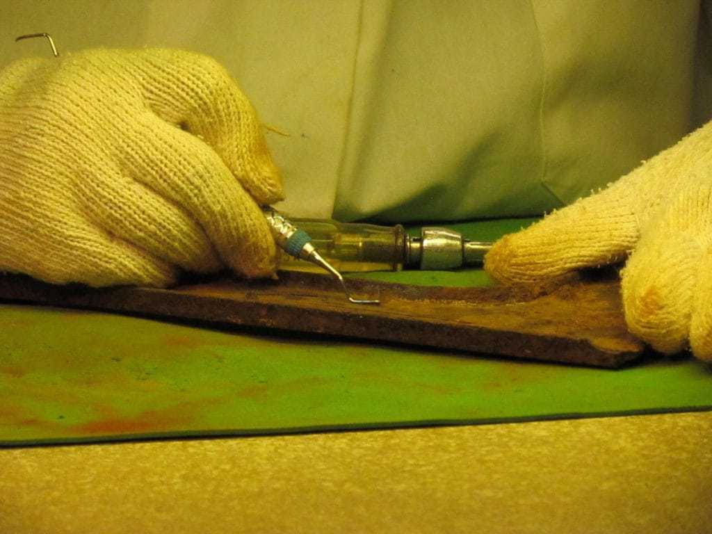 the gloved hands of a restoration worker handle a small cleaning device they use to scrape an artifact.
