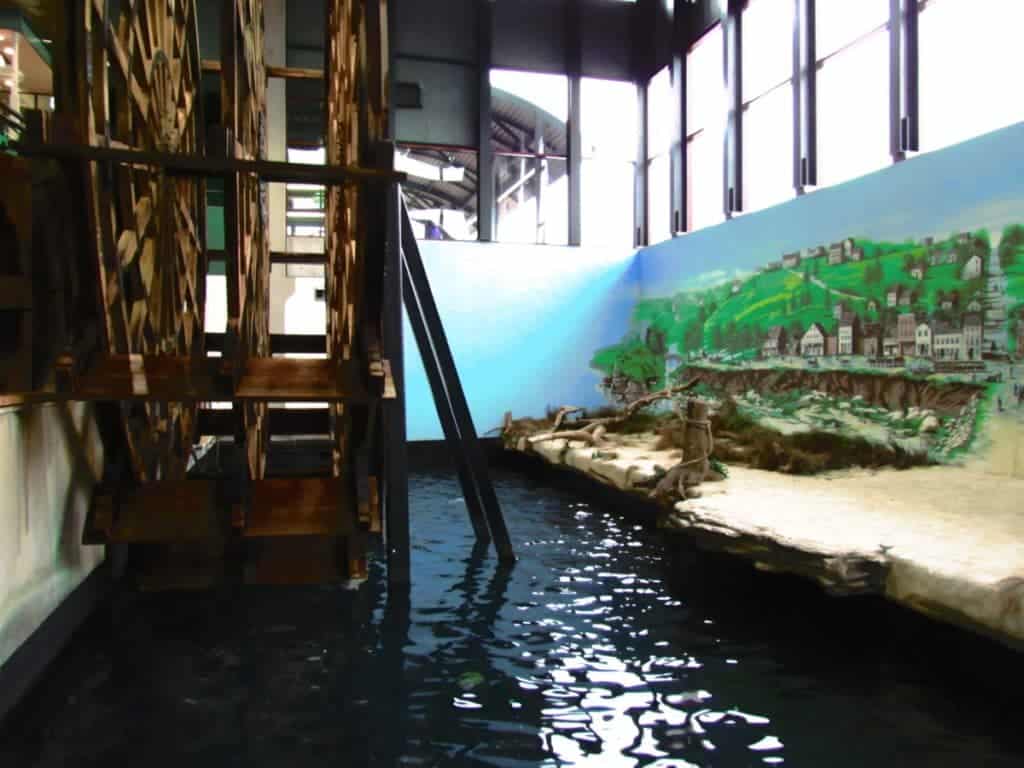 A large wooden paddle-wheel is displayed in a recreation of a riverbank scene. 