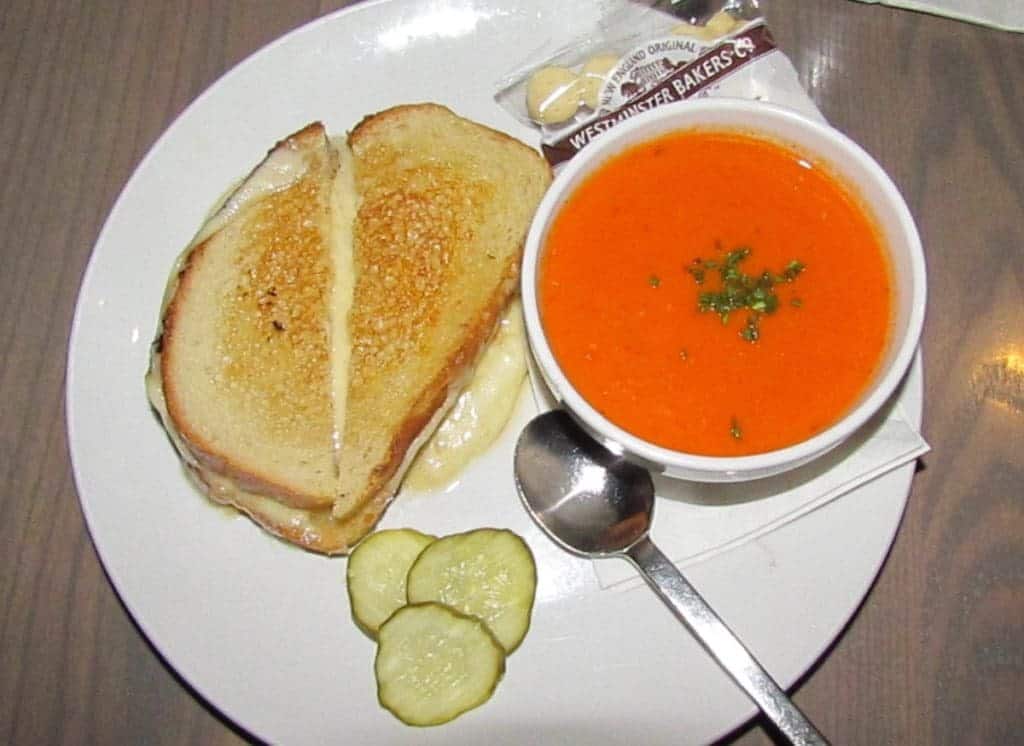A white plate holds a dinner combination of grilled cheese sandwich, picle slices, and a bowl of tomato bisque. A package of oyster crackers and a spoon rest near the bowl.