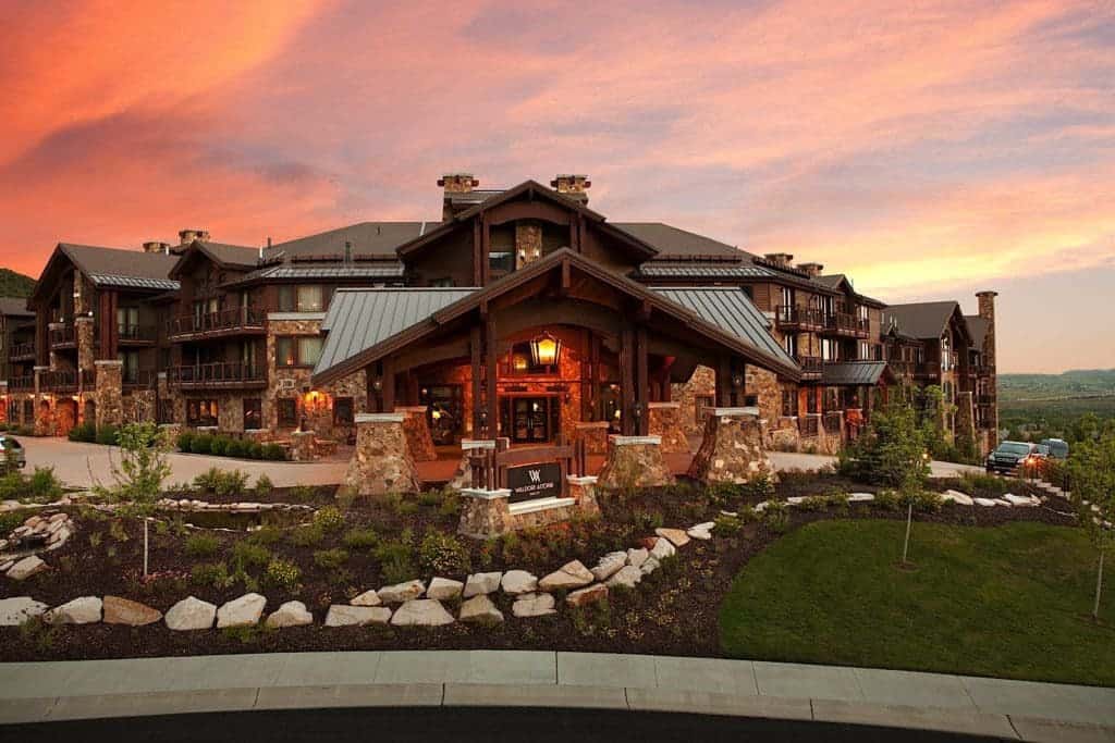 The large stone and wood lodge is the Waldorf Astoria Hotel in Park City, Utah. Rocks and greenery adorn the front lawn, as the waning sunlight casts red hues on the evening sky.
