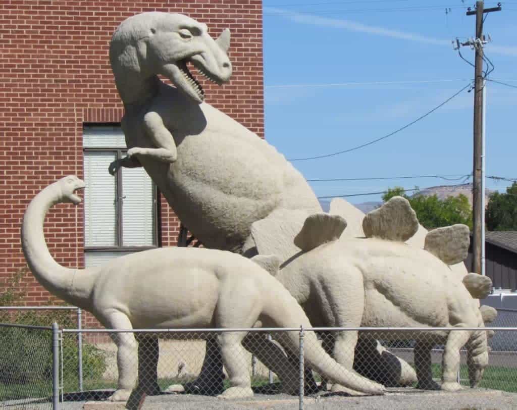 Three concrete cast dinosaur replicas are located within a chain link area in front of a brick building in Dinosaur, Colorado.