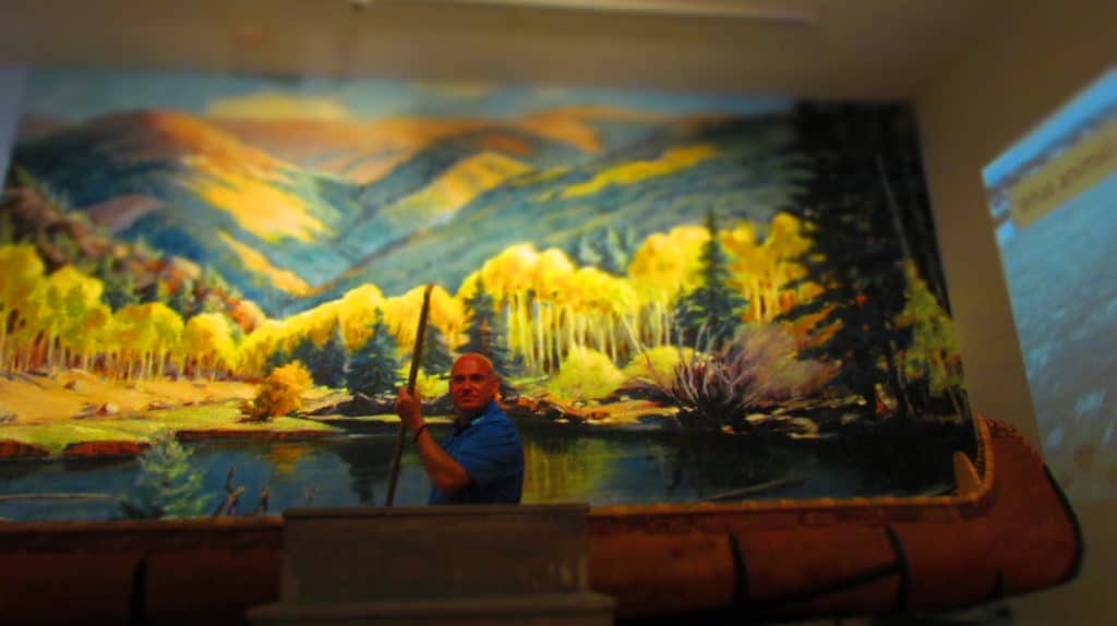 The author poses in a canoe poised in front of a scenic mural.